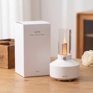 May include: A white WFM flame aroma diffuser with a 150ml capacity, featuring a cylindrical glass top, a metallic inner core emitting a warm orange light, and a circular power button on the base, accompanied by its matching white product box.