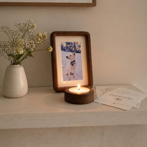 Peut inclure: Une photo encadrée en bois d'un chien, une bougie allumée dans un support en bois et un vase de fleurs blanches sont posés sur une surface blanche. La flamme de la bougie brille vivement. Plusieurs cartes blanches sont à côté de la bougie.