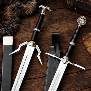 May include: Two metal swords with black wrapped handles and silver-toned crossguards. One sword features a wolf-shaped pommel, while the other has a circular emblem. Both blades are accompanied by black leather sheaths with silver accents.