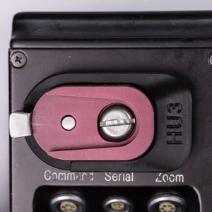 May include: Close-up of a black and red device with the text "HU3" and "Command Serial Zoom". The device has a silver metal lever and screws. The background is a light gray.