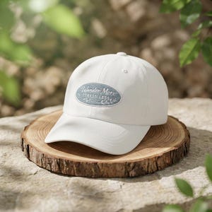 Peut inclure: Casquette de baseball blanche avec une visière incurvée. La casquette présente un écusson brodé avec les mots "Wander More Stress Less" dans un ovale bleu. Le chapeau est présenté sur une surface en bois.