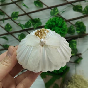 May include: A white, shell-shaped decorative item with a bow and pearl accent. The item is held in a hand, with a green and brown background. The bow is made of a sheer, light-coloured fabric.