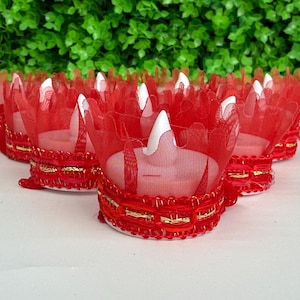 Könnte beinhalten: Eine Sammlung roter Tüll-Kerzenhalter mit goldenen Verzierungen. Jeder Halter hat eine kronenartige Form und eine weiße Kerze im Inneren. Die Halter sind auf einer weißen Oberfläche mit grünem, blättrigem Hintergrund angeordnet.