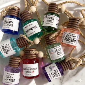 Puede incluir: Una variedad de ambientadores para coche en pequeños frascos de vidrio con tapones de madera. Cada frasco tiene un líquido de diferente color y una etiqueta con un nombre de aroma único, como "Mahogany Teakwood" y "Cactus Blossom". Los frascos están unidos por una cuerda dorada y blanca.