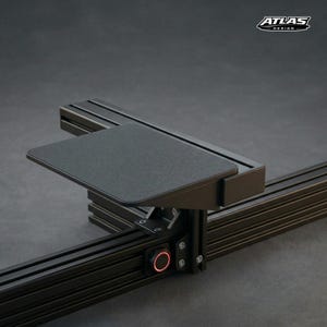 Peut inclure: Fixation de bureau en métal noir avec un petit plateau rectangulaire. La fixation comporte un bouton rouge éclairé et le logo "ATLAS DESIGN". Le design est élégant et moderne, adapté à une configuration de jeu ou de poste de travail.