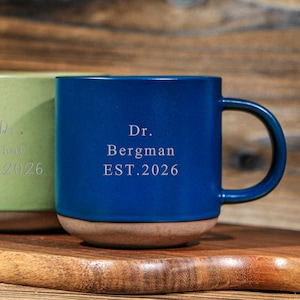 Könnte beinhalten: Eine weiße Keramik-Tasse mit schwarzem Henkel und dem Text "But First, Coffee" in schwarzer, fetter Schrift. Die Tasse steht auf einer Holzoberfläche. Die Tasse ist etwa 10 cm hoch.