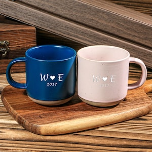 May include: Two ceramic mugs, one blue and one pink, sit on a wooden cutting board. Each mug has a white design with the letters "W E", a heart, and the year "2017". The mugs have a natural-colored base and are set against a wooden background.