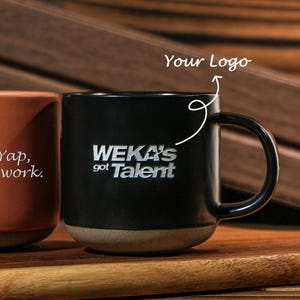 May include: A black ceramic mug with a handle, featuring the white engraved text "WEKA'S got Talent" on the front. A white arrow points to the text with the label "Your Logo" above it. The mug has a smooth black finish and a light grey base.
