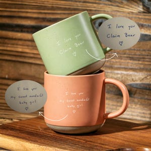 May include: A sage green ceramic mug with the text I love you Claire Bear and a heart, stacked on a peach-coloured ceramic mug with the text I love you my sweet wonderful baby girl and a heart. Both mugs feature a speckled finish and a base.