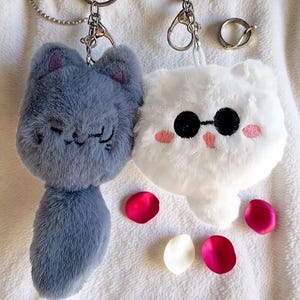 May include: Two plush cat keychains, one gray and one white, with key rings. The gray cat has embroidered details and a small tail. The white cat wears sunglasses and has pink cheeks. Scattered rose petals add a touch of color.