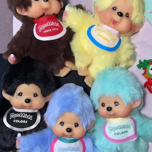 May include: A collection of Monchhichi plush dolls in various colors, including brown, yellow, black, blue, and teal. Each doll has a bib with the "Monchhichi" logo and the word "COLORS" or "SINCE 1974".
