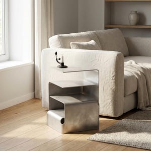 May include: A contemporary, brushed metal side table with two shelves and a curved design. A black candle holder with a lit candle sits on the top shelf. The table is next to a beige sofa in a well-lit room.