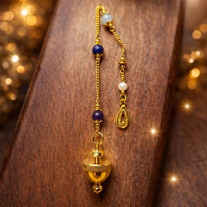 Puede incluir: Un péndulo dorado con una cadena decorativa con cuentas azules, moradas y blancas. El péndulo tiene una base en forma de campana y se muestra sobre una superficie de madera. El diseño es ornamentado y elegante.