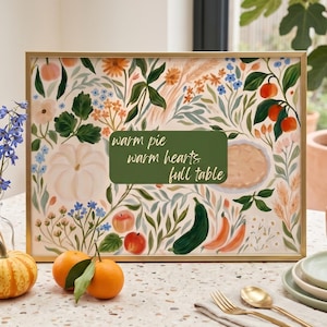 May include: A rectangular framed print featuring a botanical illustration of pumpkins, apples, flowers, and a pie. A central green rectangular box contains the white text: warm pie, warm hearts, full table. The frame is gold-toned and metallic.