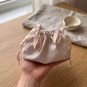 May include: A small, pink and white striped drawstring bag. The bag is made of fabric and has a gathered top with matching fabric ties. The bag is held in a hand, with a small ceramic bowl and a linen cloth in the background.