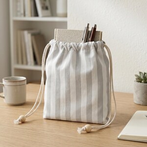 Könnte beinhalten: Ein grau-weiß gestreifter Kordelzugbeutel mit einer hellen Holzperle am Ende des Kordelzugs. Die Tasche enthält Bücher und Stifte. Eine Tasse und eine Sukkulente in einem Betontopf stehen ebenfalls auf dem Tisch.