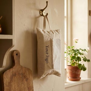 Peut inclure: Un sac en lin beige avec le texte "Your logo" suspendu à un crochet en laiton sur un mur crème. Une planche à découper en bois et un petit pot marron sont également visibles. Une plante en pot est sur un rebord de fenêtre.