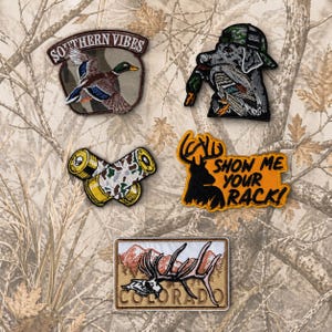 May include: A collection of embroidered patches with hunting and outdoor themes. Designs include a mallard duck, a dog with a duck, crossed shotgun shells, a deer silhouette with text, and a Colorado-themed patch with a deer skull.