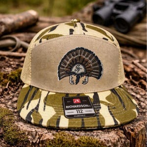 Op de afbeelding: Een camouflage trucker pet met een bruin voorpaneel en een mesh achterkant. De pet heeft een geborduurd kalkoenontwerp en het Richardson-merklabel. De klep en het voorpaneel hebben een camouflagepatroon.