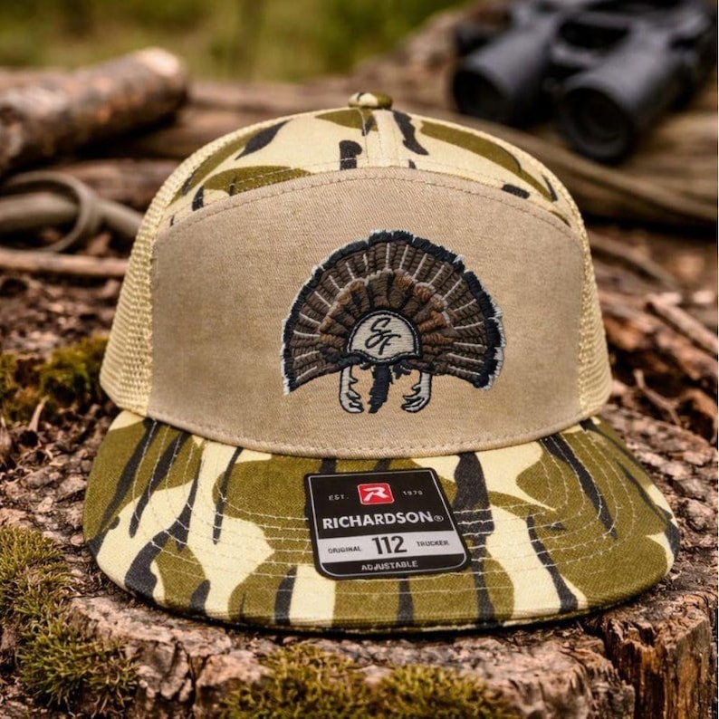 Op de afbeelding: Een camouflage trucker pet met een bruin voorpaneel en een mesh achterkant. De pet heeft een geborduurd kalkoenontwerp en het Richardson-merklabel. De klep en het voorpaneel hebben een camouflagepatroon.
