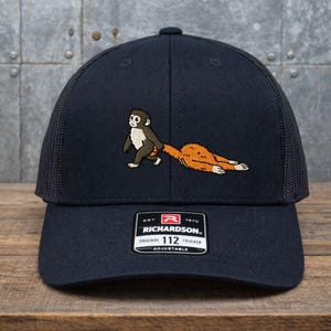 May include: Navy blue trucker hat with a mesh back and a curved brim. The hat features an embroidered design of a monkey pulling a long, orange tail. The Richardson brand label is visible on the front.