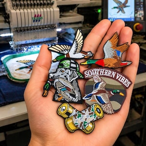 May include: A hand holds several embroidered patches featuring ducks in flight and other duck-related designs. The patches are in various colors, including green, brown, and white. One patch has the text "SOUTHERN VIBES".