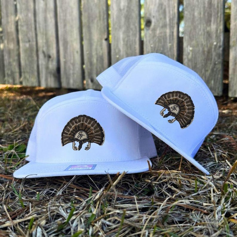 Op de afbeelding: Twee witte truckerpetten met een bruin kalkoenontwerp geborduurd op de voorkant. De petten hebben een platte klep en een mesh achterkant. Het kalkoenontwerp heeft een bruin lichaam en een waaier van veren.
