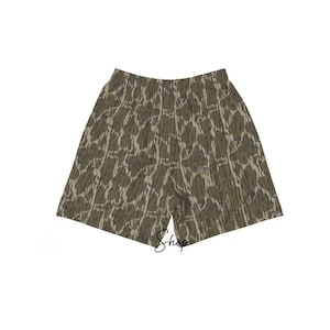 May include: A pair of shorts with a camouflage pattern in shades of green, brown, and tan. The shorts have an elastic waistband and a relaxed fit. The word "Shop" is written in cursive on the shorts.
