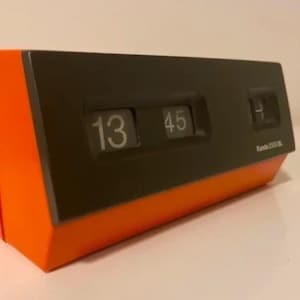 May include: An orange and black flip clock displaying the time 13:45. The clock has a rectangular shape with a black face and orange sides. The brand name "Randa 2000" is visible on the clock face.
