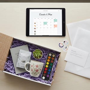 May include: An open gift box filled with purple crinkle paper, a floral mug, a small succulent, a watercolor palette, and a book. A tablet displays a calendar with the words "Curate & Plan". Stickers and a welcome guide are also present.