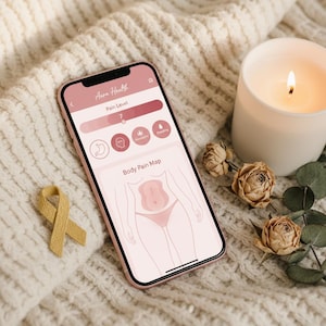Può includere: Uno smartphone che mostra l'app "Aura Health" con una mappa del dolore corporeo e un indicatore del livello di dolore. Una candela accesa, rose secche e un nastro giallo sono accanto al telefono su una superficie bianca strutturata.