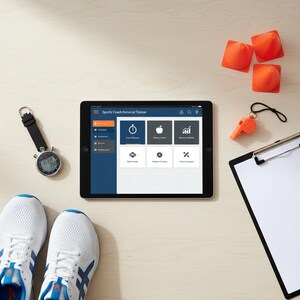 Op de afbeelding: Een bovenaanzicht van sporttrainingsapparatuur op een licht houten oppervlak. De afbeelding toont een tablet met "Sports Coach Personal Trainer", witte sportschoenen, een stopwatch, oranje kegels, een fluitje en een klembord.