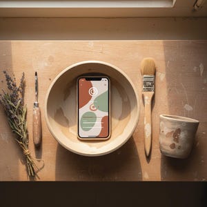 May include: A wooden bowl holds a smartphone with a colorful abstract design. A paintbrush, pottery tool, small ceramic cup, and a bundle of lavender are arranged on a wooden surface. The phone's screen displays a design with orange, green, and white shapes.