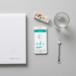 Peut inclure: Un smartphone blanc affichant une application médicale avec un graphique, à côté d'un pilulier, d'un verre d'eau, d'un dossier blanc avec le texte "CONSENT FORMS" et d'un bracelet bleu et blanc. Les objets sont disposés sur une surface grise.