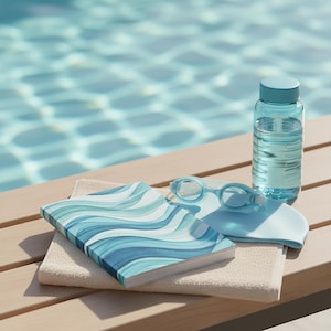 Peut inclure: Une bouteille d'eau bleu clair, des lunettes de natation et un bonnet de bain sont posés à côté d'un carnet avec un motif de vagues bleues et blanches sur une serviette beige. Les articles sont disposés sur une surface en bois avec une piscine en arrière-plan.