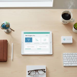 May include: A workspace scene with a tablet showing "CRM Navigator Pro", a coffee cup, a small globe, a notebook, a stylus, a business card, a keyboard, a book, and eyeglasses. The items are arranged on a light wood surface.