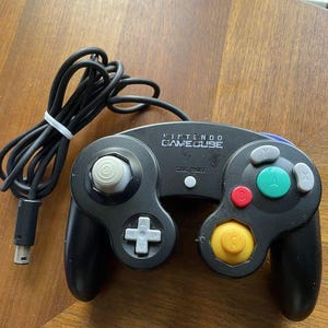 May include: Black Nintendo GameCube controller with a variety of colored buttons, including red, green, and yellow. The controller has a white analog stick and a black cable.