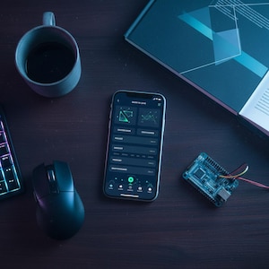 Op de afbeelding: Een donkerhouten bureau met een smartphone waarop een app-interface wordt weergegeven. Een kop koffie, een boek, een toetsenbord, een muis en een printplaat liggen ook op het bureau. De app-interface heeft groene geometrische vormen en tekst.