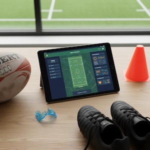 Peut inclure: Une tablette affichant des analyses de match est posée sur une surface en bois, à côté d'un ballon de rugby, d'un cône orange, d'un protège-dents bleu et de crampons noirs. La tablette montre un schéma de terrain vert et des données.