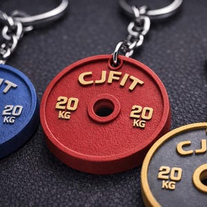 Peut inclure: Gros plan sur trois porte-clés en forme de disques de poids, bleus, rouges et dorés. Chaque porte-clé porte l'inscription "CJFIT" et "20 KG". Les porte-clés sont attachés à des chaînes argentées.