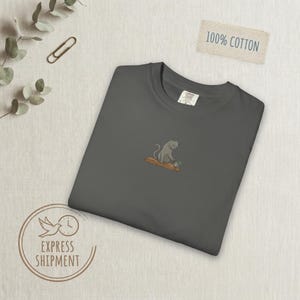 Könnte beinhalten: Dunkelgraues T-Shirt mit einem gestickten Design einer grauen Katze, die auf einem Baumstamm mit Blumen sitzt. Das Shirt hat ein Etikett mit der Aufschrift "Comfort Colors". Ein kleines Etikett lautet "100% Baumwolle". Das Shirt ist sauber gefaltet.