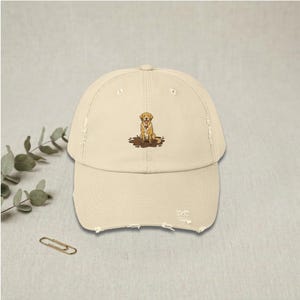 Puede incluir: Gorra de béisbol color crema desgastada con una ilustración bordada de un golden retriever sentado en el barro. La gorra tiene una visera curva y una correa ajustable, perfecta para amantes de los perros.