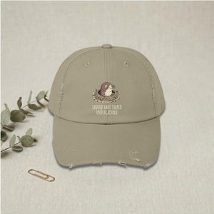 May include: A distressed, khaki-colored baseball cap featuring an illustration of a mole peeking out of a burrow. The text "SHOULD HAVE STAYED UNDERGROUND" is printed below the graphic. The cap has a curved brim and a worn appearance.