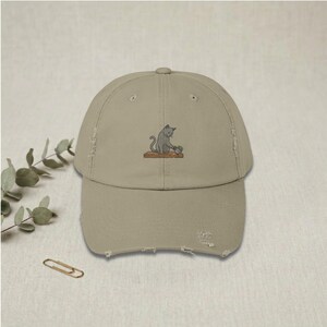 Könnte beinhalten: Beige Baseballkappe mit Used-Look und einem gestickten Design einer grauen Katze, die sich um eine kleine Topfpflanze kümmert. Die Kappe hat einen gebogenen Schirm und einen abgenutzten, Vintage-Look mit ausgefransten Kanten.
