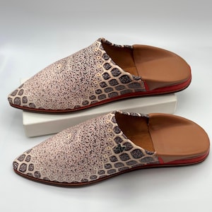Puede incluir: Un par de zapatillas de estilo marroquí. Las zapatillas presentan un diseño estampado en tonos beige y marrón en la parte superior, con un interior y suela de cuero marrón. La suela tiene un ribete rojo. Las zapatillas se exhiben sobre una superficie blanca.