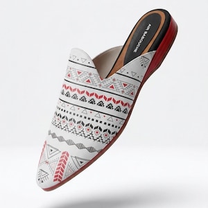 Puede incluir: Zapato blanco sin cordones con un estampado geométrico en rojo, negro y blanco. El zapato tiene una suela roja y una plantilla negra. El texto "AIR BABOUCHE" está impreso en la plantilla. Talla 39.