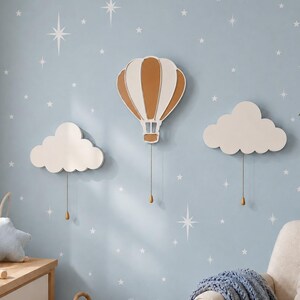 May include: Three wall-mounted decorations featuring a central hot air balloon in white and tan, flanked by two white cloud shapes. Each piece includes a thin cord hanging downward with a small, matching teardrop-shaped weight at the bottom.