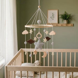 May include: A wooden baby mobile with a circular frame, featuring hanging wooden stars, clouds, and a crescent moon. The mobile also includes gray crocheted elements and a large gray star. The mobile hangs above a wooden crib.