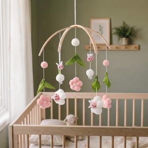May include: A hanging nursery mobile featuring a curved wooden frame with suspended felt elements including pink flowers, white clouds, green leaves, and small pink and white bees. The decorative items are attached to the frame with white strings.
