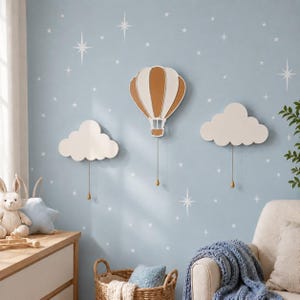 May include: A wall-mounted decor set featuring a central hot air balloon in white and tan, flanked by two white cloud shapes. Each piece includes a thin vertical cord with a small teardrop-shaped weight hanging from the bottom edge.
